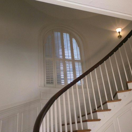 White plantation shutters adorning arched window located in spiral stairwell.